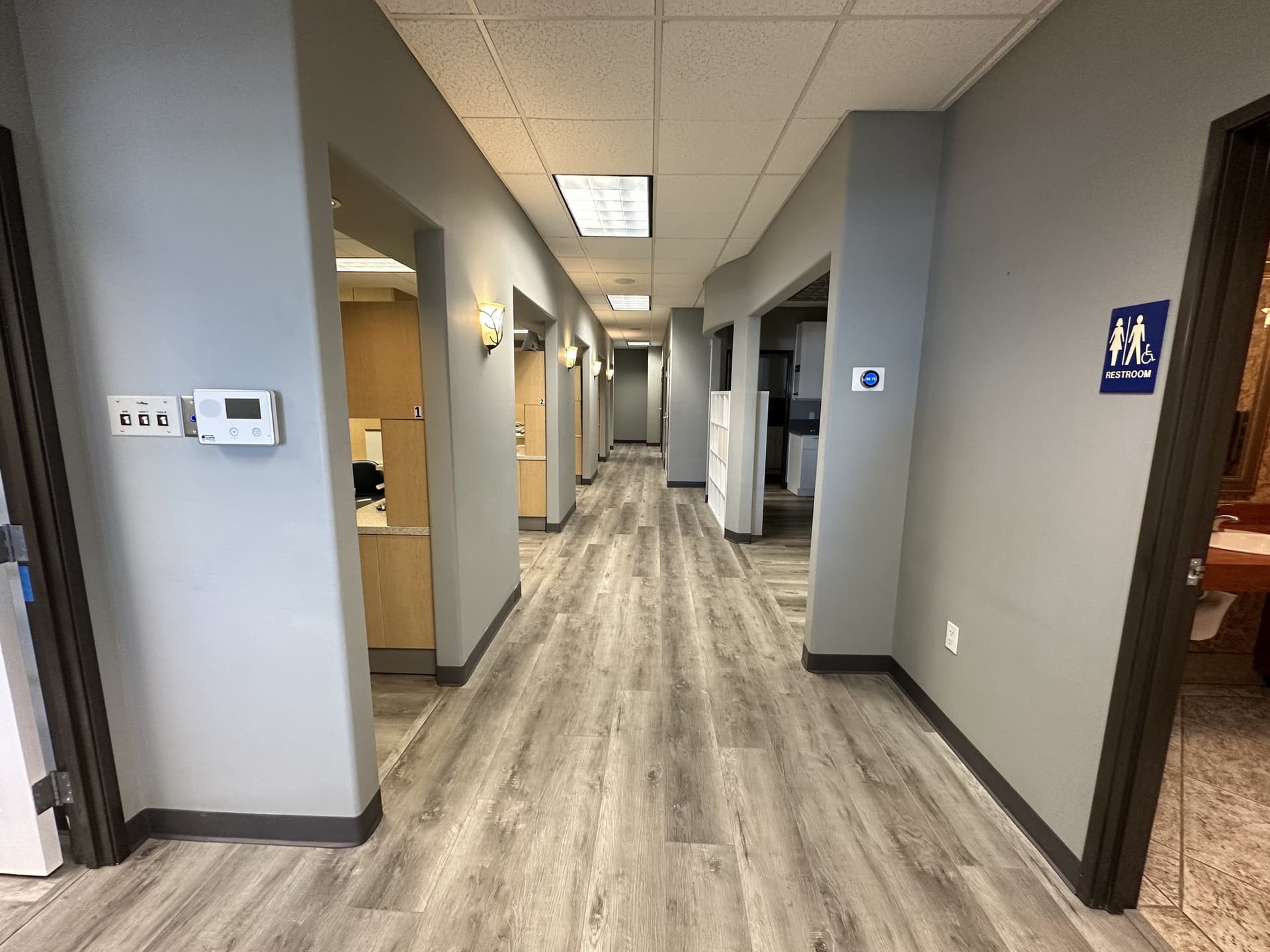 Looking down the gray office hallway, with open operatory bays on the left and a restroom door on the right.