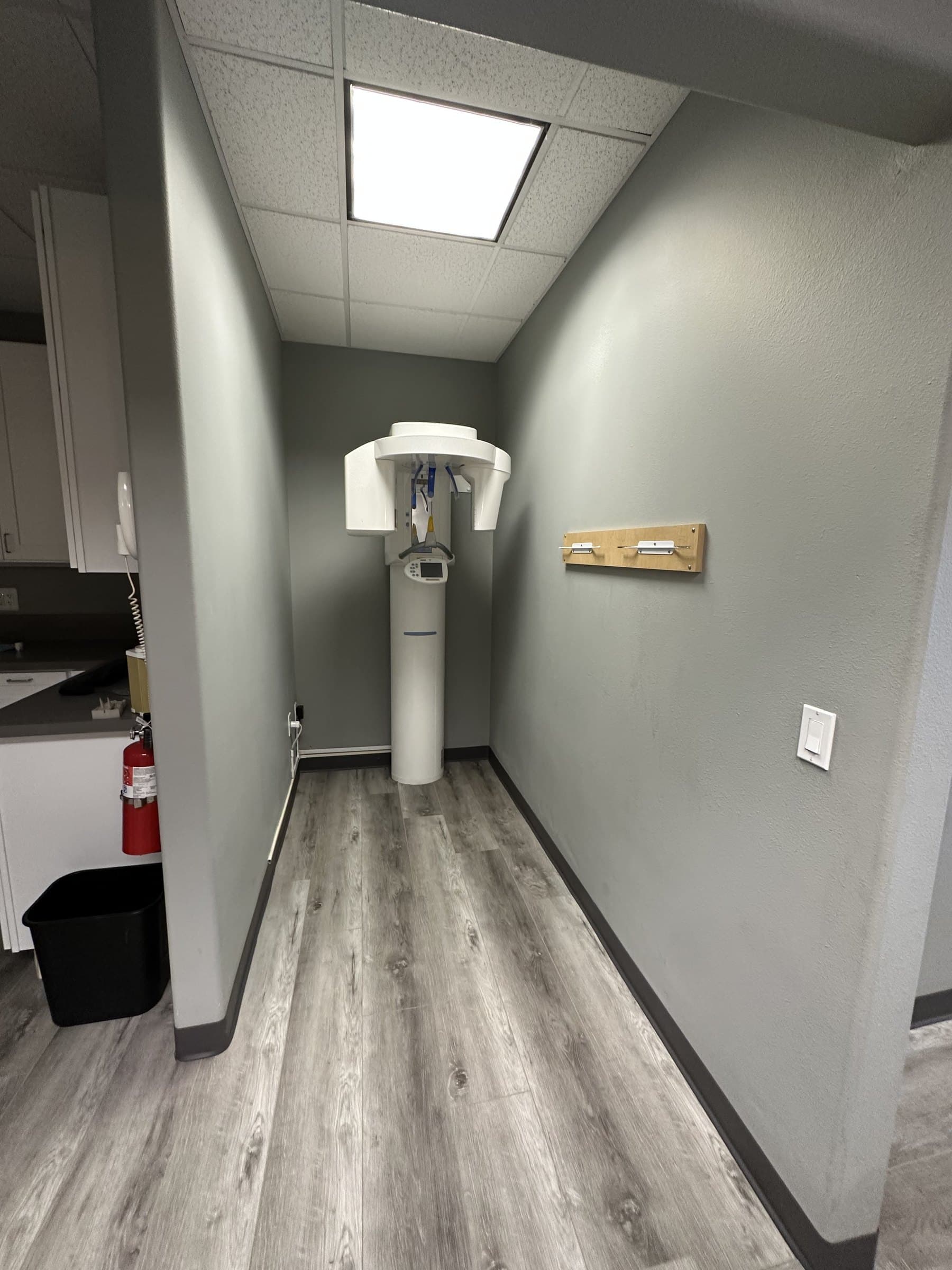 Upright CBCT and panoramic X-ray machine standing in a gray alcove at the end of a short hall.