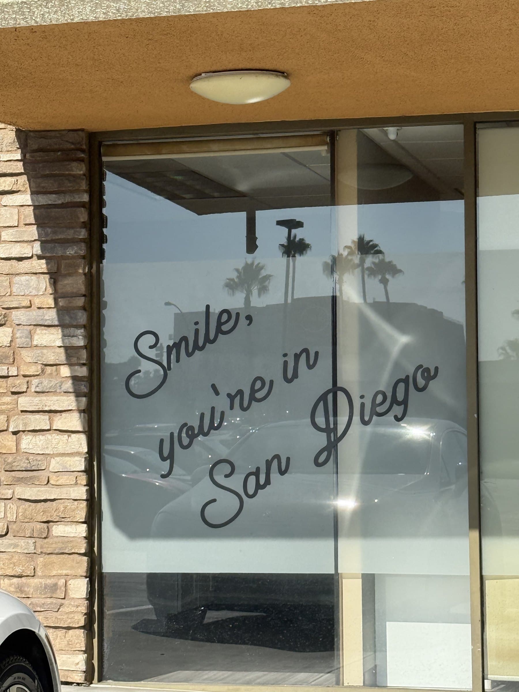 Close photograph of the front window vinyl in script that reads Smile, you're in San Diego, with palm trees reflected in the glass.